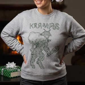 Op de afbeelding: Een grijs gemêleerd sweatshirt met ronde hals en een donkergroene lijntekening van Krampus die een zak draagt en een knots vasthoudt. Het woord KRAMPUS staat in een gestileerd lettertype boven de figuur, die uit complexe, verweven lijnen bestaat.