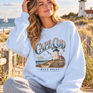 Könnte beinhalten: Ein hellblaues Sweatshirt mit Rundhalsausschnitt und einem Grafikdruck eines Leuchtturms auf einer Sanddüne mit dem Text CAPE COD, RACE POINT und PROVINCETOWN MASSACHUSETTS. Das Motiv zeigt eine fliegende Möwe, Wolken und ein Segelboot.