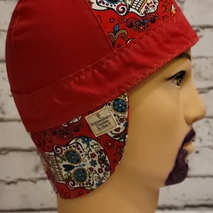 Può includere: Cappello da saldatura rosso con un motivo a teschi e fiori. Il cappello ha una patta che può essere indossata verso il basso per proteggere il collo e le orecchie. La patta ha un'etichetta che recita "Extraordinary Creations by Toni T."