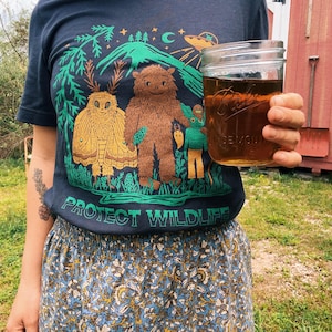 May include: Navy blue t-shirt with a graphic featuring a brown Bigfoot-like creature, a moth, and a green creature, with the text "PROTECT WILDLIFE". A clear glass jar with a brown liquid is held in the hand.