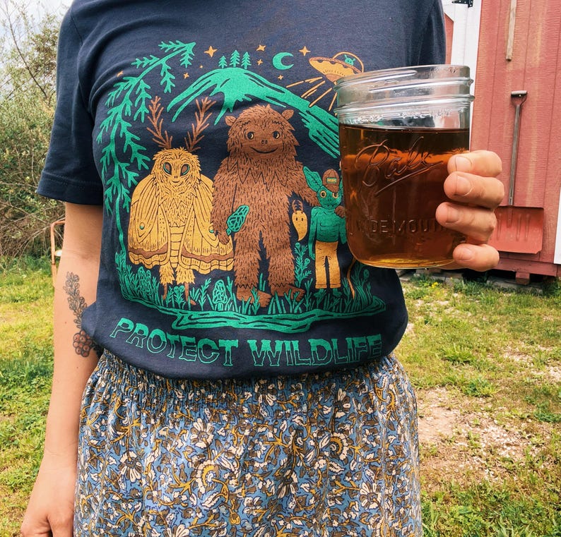 May include: Navy blue t-shirt with a graphic featuring a brown Bigfoot-like creature, a moth, and a green creature, with the text "PROTECT WILDLIFE". A clear glass jar with a brown liquid is held in the hand.