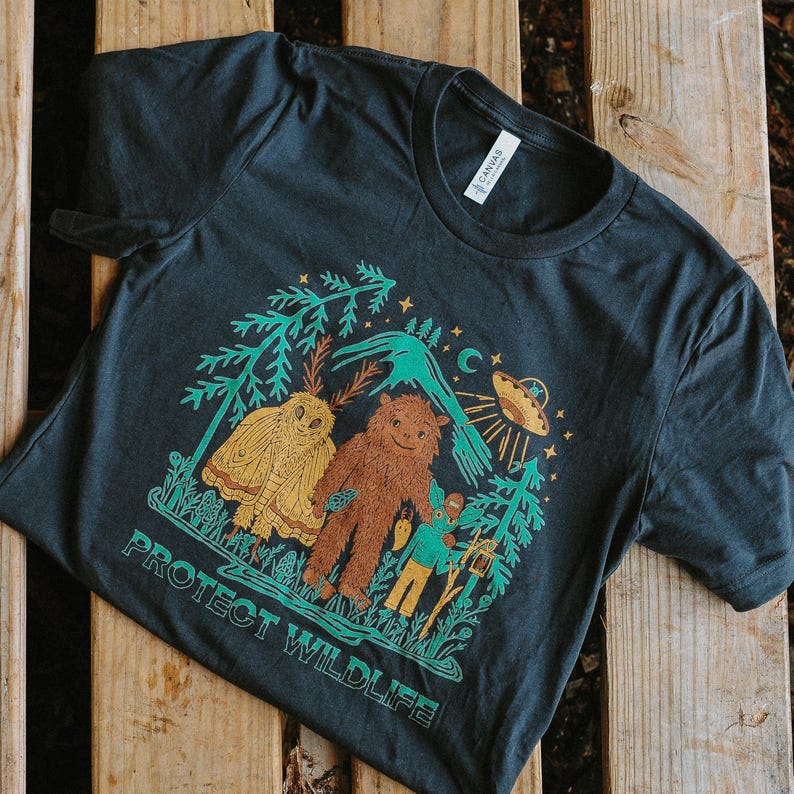 May include: Dark gray t-shirt with a colorful graphic featuring an owl, Bigfoot, and a figure with a staff, framed by trees and foliage. The text "PROTECT WILDLIFE" is printed below the graphic.