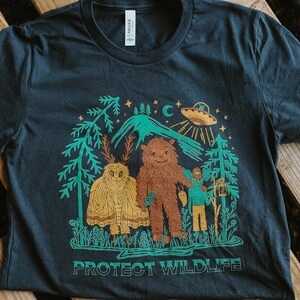 May include: Dark gray t-shirt featuring a whimsical illustration with a brown Bigfoot, a yellow moth, and a teal alien. The design includes trees, a mountain, and a UFO, with the text "PROTECT WILDLIFE" in green.