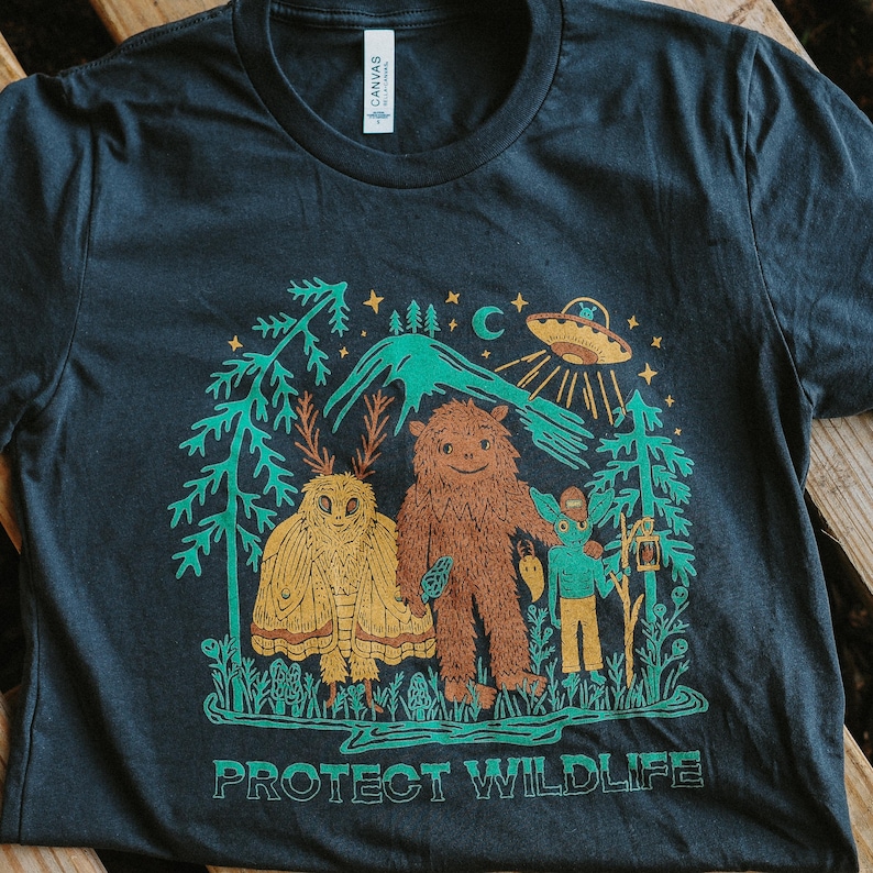 May include: Dark gray t-shirt featuring a whimsical illustration with a brown Bigfoot, a yellow moth, and a teal alien. The design includes trees, a mountain, and a UFO, with the text "PROTECT WILDLIFE" in green.