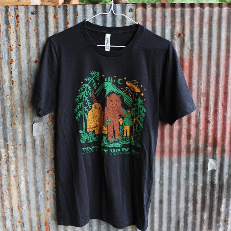 May include: Black t-shirt with a colorful graphic featuring a Bigfoot-like creature, an owl, and a person in a forest setting. The design includes a mountain, a crescent moon, and a UFO. The text "PROTECT WILDLIFE" is printed at the bottom.