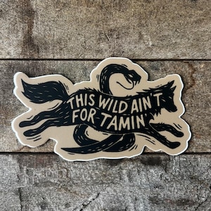 May include: A sticker with a black wolf and snake design. The wolf is leaping, and the snake is coiled around it. The text "THIS WILD AIN'T FOR TAMIN'" is written across the center in black. The sticker has a tan background and a white border.