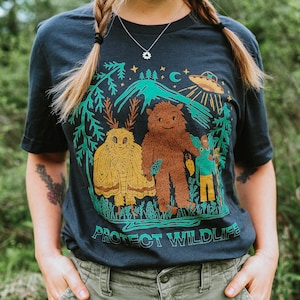 Puede incluir: Camiseta azul marino oscuro con un gráfico colorido que presenta una polilla, Bigfoot y una criatura con un bastón, ambientada en una escena de montaña y bosque. El texto "PROTECT WILDLIFE" está impreso en la parte inferior.