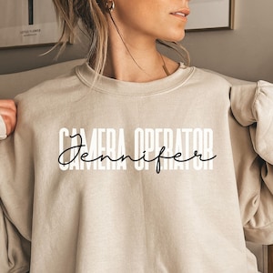 May include: A beige sweatshirt with the text "CAMERA OPERATOR Jennifer" printed in black.