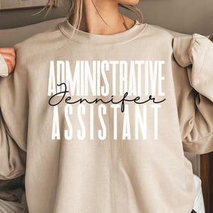 May include: A beige sweatshirt with the words "ADMINISTRATIVE ASSISTANT" in white block letters. The name "Jennifer" is written in a cursive font between the words. The sweatshirt has long sleeves.