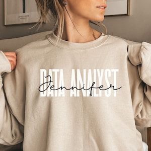Może przedstawiać: Beżowa bluza z okrągłym dekoltem z napisem "DATA ANALYST" pogrubioną czcionką bezszeryfową i "Jennifer" kursywą. Bluza ma neutralny kolor, a tekst jest biały. Zdjęcie przedstawia osobę noszącą bluzę.