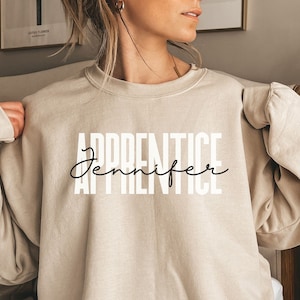 May include: A beige sweatshirt with the text "APPRENTICE Jennifer" printed in black on the front.