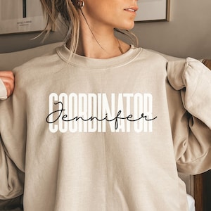 Könnte beinhalten: Ein beiger Pullover mit dem Aufdruck "COORDINATOR Jennifer" in Schwarz und Weiß.