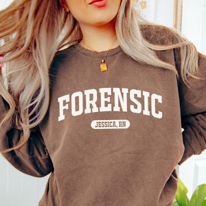 Könnte beinhalten: Ein braunes Sweatshirt mit dem Wort "FORENSIC" in weiß auf der Vorderseite bedruckt. Unter dem Wort befindet sich ein weißes Oval mit dem Text "JESSICA, RN" darin.