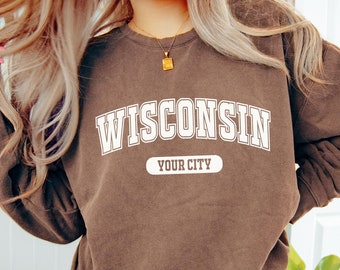 May include: A brown sweatshirt with the word "WISCONSIN" in white block letters arched across the chest, with "YOUR CITY" below. The sweatshirt is worn with black leggings.