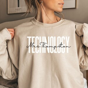 Peut inclure: Un sweat-shirt beige avec le mot "TECHNOLOGY" en grandes lettres blanches et "Mrs. Thompson" en écriture cursive. Le sweat-shirt à manches longues est de couleur neutre.