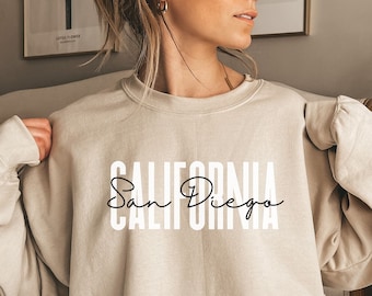 以下が含まれることがあります： 前面に「CALIFORNIA San Diego」という文字が白でプリントされたベージュのトレーナーです。