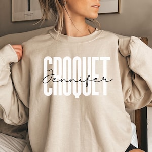 Könnte beinhalten: Ein beiger Pullover mit dem Wort "CROQUET" in weißen Buchstaben und dem Namen "Jennifer" darunter in Kursivschrift.