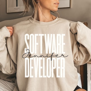 May include: Beige sweatshirt with the text "SOFTWARE DEVELOPER Jennifer" in white and black. The sweatshirt is a neutral color and has long sleeves. The text is stacked vertically, with "Jennifer" in a cursive font.