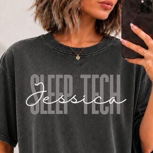 May include: A charcoal grey short-sleeve t-shirt with a vintage wash finish. The shirt features the text "SLEEP TECH" in a bold, light grey block font, overlaid with the name "Jessica" in a white, cursive script font across the center.