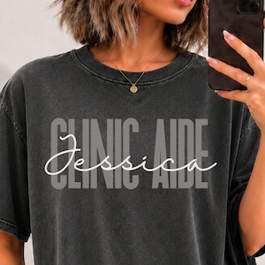 May include: A charcoal grey short-sleeve t-shirt with a vintage wash finish. The front features the text CLINIC AIDE in a bold, block font with the name Jessica written in a white cursive script layered over the centre of the text.