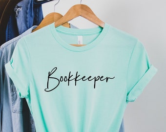 May include: A light teal t-shirt with the word "Bookkeeper" in black script. The shirt is hanging on a wooden hanger, alongside a denim shirt and a brown hat. The t-shirt is knotted at the waist.