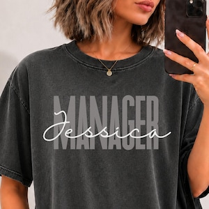 May include: A charcoal grey short-sleeve t-shirt with a vintage-washed finish. The front features the word MANAGER in large, bold, light grey block letters, overlaid with the name Jessica written in a white, flowing, cursive script font.