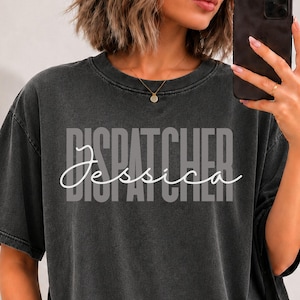 May include: A charcoal grey short-sleeve t-shirt with a vintage wash finish. The front features the word DISPATCHER in large, light grey block letters, overlaid with the name Jessica written in a white, flowing cursive script font.