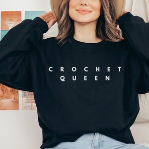 May include: Black crewneck sweatshirt with the text "CROCHET QUEEN" in white block letters. The sweatshirt is a solid black color and has long sleeves. The person in the image is wearing a tan hat.