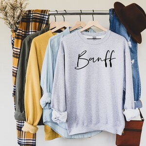 以下が含まれることがあります： 黒い筆記体で「Banff」と書かれた灰色のスウェットシャツ。スウェットシャツは、他の衣料品と一緒にラックにかけられています。