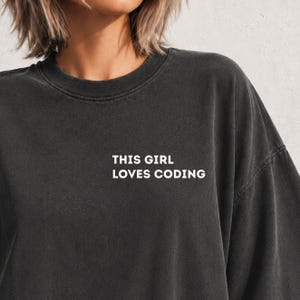 May include: A charcoal grey crewneck t-shirt with a washed-out finish. The chest features white text that reads THIS GIRL LOVES CODING in a clean, sans-serif font. The garment has a relaxed fit with dropped shoulders and a ribbed neckline.