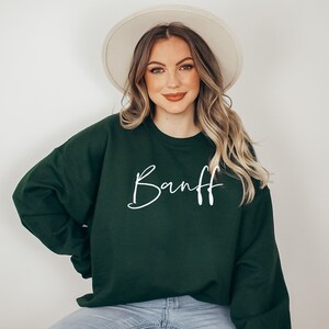 以下が含まれることがあります： 白い文字で「Banff」と書かれた濃い緑色のスウェットシャツを着た女性。彼女は薄い茶色のフェドラハットと青いジーンズも履いています。