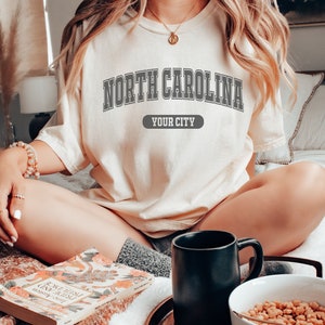K&ouml;nnte beinhalten: Cremefarbenes T-Shirt mit dem Text "NORTH CAROLINA" in grauen Blockbuchstaben. Darunter befindet sich ein graues Banner mit der Aufschrift "YOUR CITY". Das Shirt wird von einer Person getragen, die auf einem Bett mit einem Buch, einer Tasse und einer Sch&uuml;ssel M&uuml;sli sitzt.