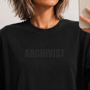 May include: A black crewneck sweatshirt with a washed, faded finish. The word ARCHIVIST is printed across the chest in a dark, tonal font that blends into the fabric. The garment features a ribbed collar and long sleeves in a solid black color.