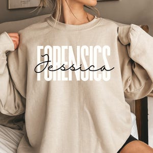Könnte beinhalten: Beigefarbenes Sweatshirt mit Rundhalsausschnitt und dem Wort "FORENSICS" in großen, weißen Blockbuchstaben. Der Name "Jessica" ist in kursiver Schrift unter dem Wort "FORENSICS" geschrieben. Das Sweatshirt hat lange Ärmel.