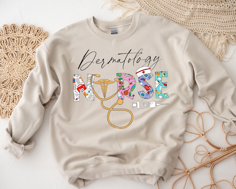May include: A beige sweatshirt with the text "Dermatology Nurse" in black lettering. The letters are decorated with colorful medical icons, including a stethoscope, a syringe, and a bandage.