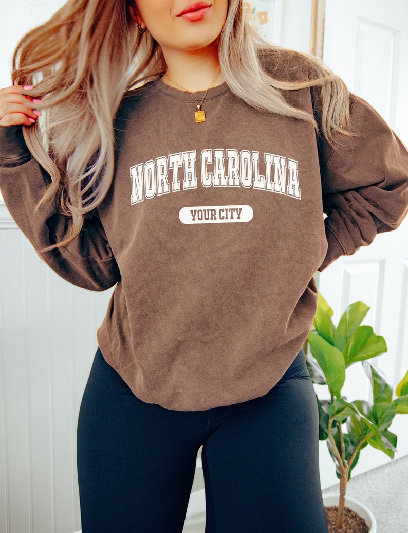 K&ouml;nnte beinhalten: Ein braunes Sweatshirt mit dem Text "NORTH CAROLINA" in wei&szlig;en Blockbuchstaben. Darunter steht der Text "YOUR CITY" in einem wei&szlig;en Rechteck. Das Sweatshirt wird mit schwarzen Leggings getragen.