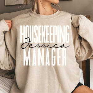 May include: Beige sweatshirt with the words "HOUSEKEEPING Jessica MANAGER" printed in white and black. The text is centered on the front of the sweatshirt. The sweatshirt has long sleeves and a crew neck.