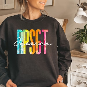 Può includere: Felpa nera con un design ad acquerello arcobaleno che scrive "RPSCT" e il nome "Jessica" in bianco.