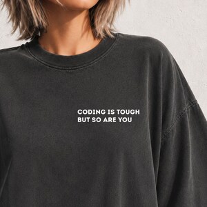 May include: A charcoal grey crewneck t-shirt featuring the white text "CODING IS TOUGH BUT SO ARE YOU" printed on the left chest area. The garment has a relaxed fit with dropped shoulders and a classic ribbed neckline, made of a washed fabric.