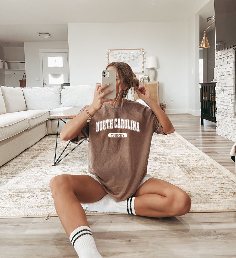 K&ouml;nnte beinhalten: Ein braunes T-Shirt mit dem Text "NORTH CAROLINA YOUR CITY" in wei&szlig;en Blockbuchstaben. Das Shirt wird mit wei&szlig;en Shorts und wei&szlig;en Socken mit schwarzen Streifen getragen. Die Person sitzt auf einem hellfarbigen Teppich.