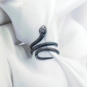May include: A dark grey snake ring coiled around white fabric. The ring has a detailed snake head and body, wrapping around the finger. The ring is made of a dark metal.