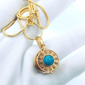 Könnte beinhalten: Eine goldfarbene Halskette mit einem runden Anhänger mit einem türkisfarbenen Stein. Der Anhänger hat ein dekoratives Blumenmuster um den Stein. Die Kette hat ein dünnes, gedrehtes Design. Die Halskette wird auf einer weißen Oberfläche präsentiert.