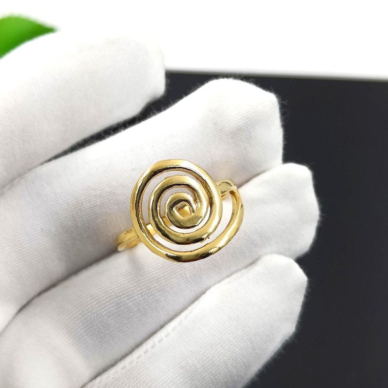 May include: A gold-colored ring featuring a spiral design. The ring is held against a white background, showcasing its intricate, openwork pattern. The ring's design resembles a nautilus shell.