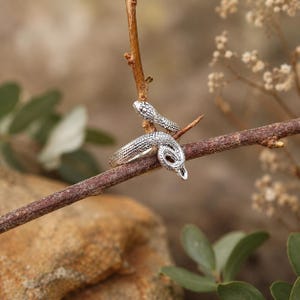 May include: A silver-coloured metal ring shaped like a coiled snake with detailed scale textures. The ring features an open design where the snake's head and tail overlap, wrapping around a thin brown twig in a natural outdoor setting.