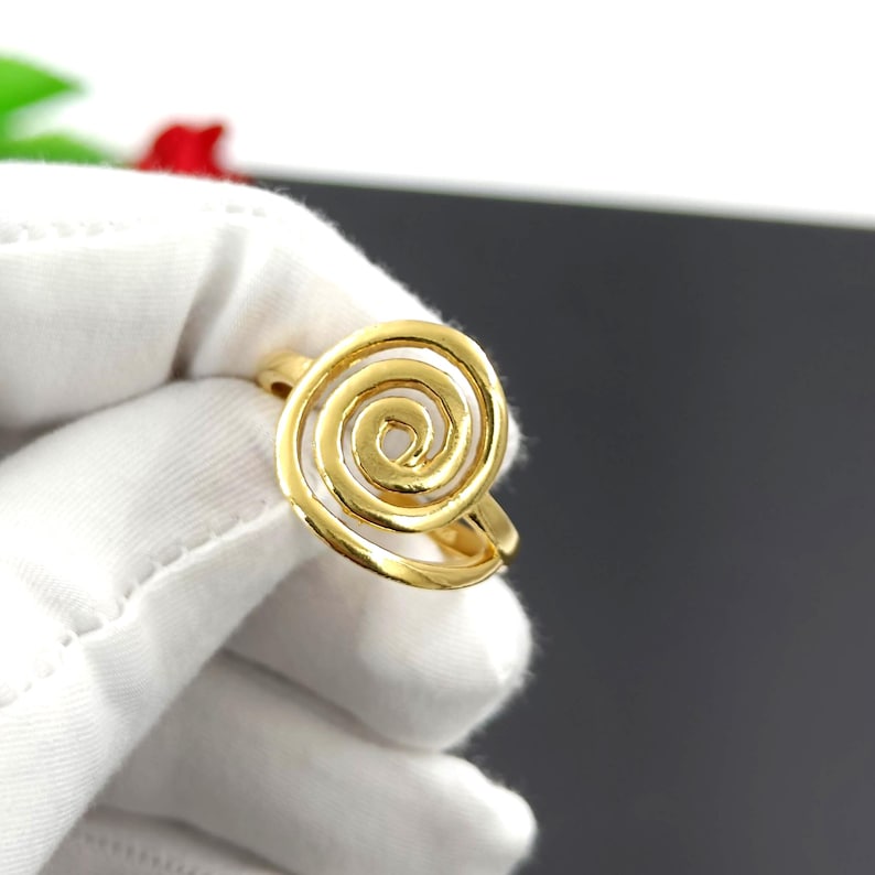 May include: A gold-colored ring featuring a spiral design. The ring's spiral is open, creating a three-dimensional effect. The ring is held against a white background, highlighting its bright, metallic finish. The ring is a piece of jewelry.