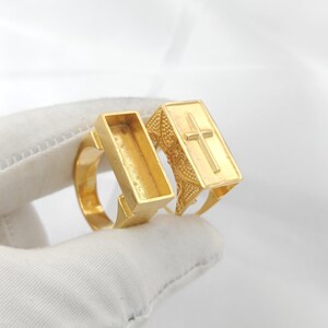 Op de afbeelding: Twee goudkleurige ringen. Eén ring heeft een rechthoekige bovenkant met een open binnenkant. De andere ring heeft een rechthoekige bovenkant met een kruisontwerp. De ringen staan tegen een witte achtergrond.