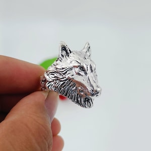 Op de afbeelding: Een zilverkleurige ring met een gedetailleerd wolfskopontwerp. De wolfskop is gebeeldhouwd met ingewikkelde vacht en gelaatstrekken. De ring wordt tegen een lichte achtergrond gehouden, wat de metaalglans benadrukt.