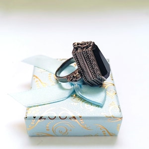 Puede incluir: Un anillo negro con una gran piedra negra facetada engastada en un engaste adornado plateado. El anillo se muestra en una caja de regalo azul claro con un patrón dorado.