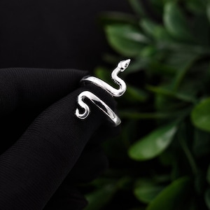 Puede incluir: Un anillo de tono plateado con forma de serpiente enroscada. La banda de metal presenta un acabado liso y pulido con una cabeza detallada y una cola curva que se envuelve alrededor del dedo. El diseño es abierto, permitiendo un ajuste.