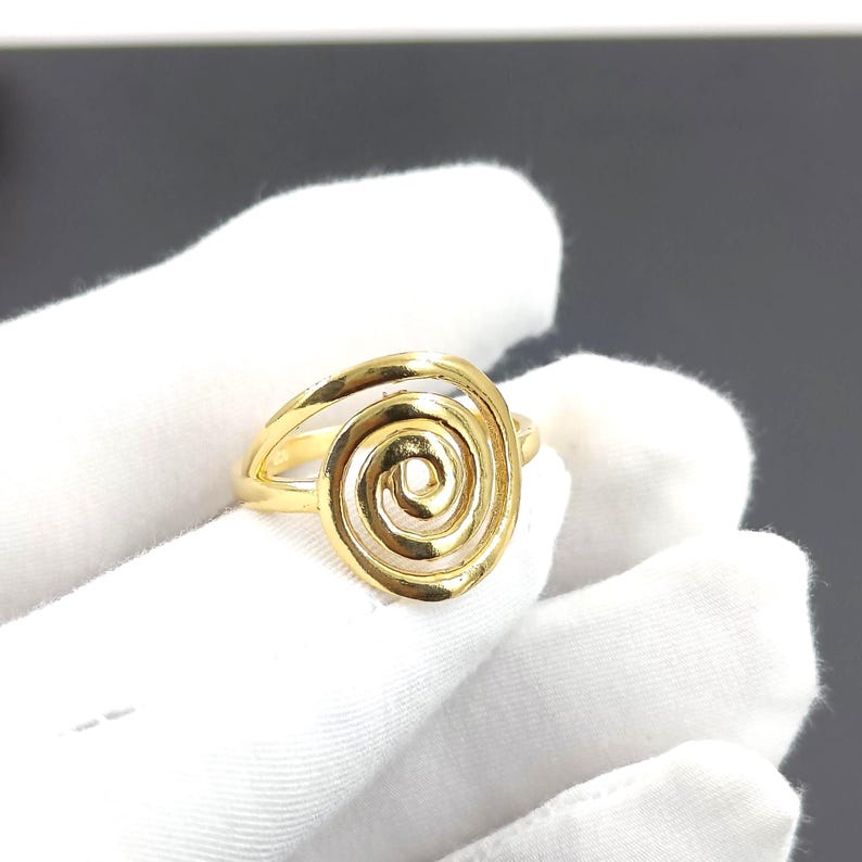 May include: A gold-colored ring featuring a spiral design. The ring has a band and a central spiral element, creating an open, airy aesthetic. The ring is held against a white background.